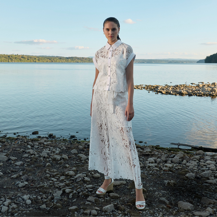 Woman in a white lace dress standing on a rocky shoreline with water and sky in the background